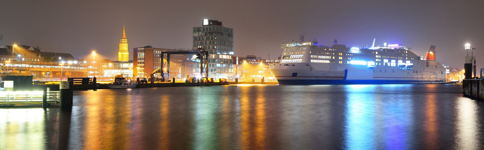 Stort, hvidt krydstogtskib i den oplyste passagerhavn om natten. Panoramisk bybillede. Kiel, Tyskland