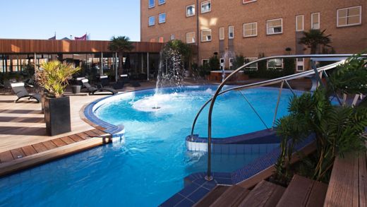 Pool med en vattenfontän och solstolar med utsikt över ett hotell.