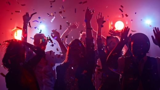 Two young women dance and laugh together at a lively party. They are surrounded by shimmering confetti and metallic streamers under vibrant pink and purple lights. One woman wears a sparkling sequin dress, while the other cheers with her hands raised. The