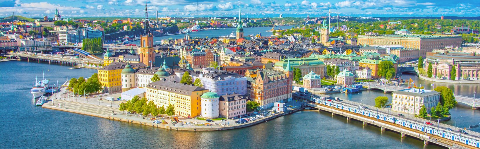 A panoramic view of bridges and buildings in central Stockholm with its mixture of traditional modern buildings, tall spires and all surrounded by water and low hills on a sunny day.