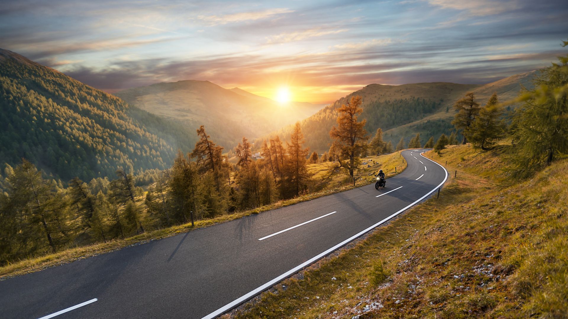 A beautiful valley landscape showing a rising sun and an open road with a single motorcyclist.