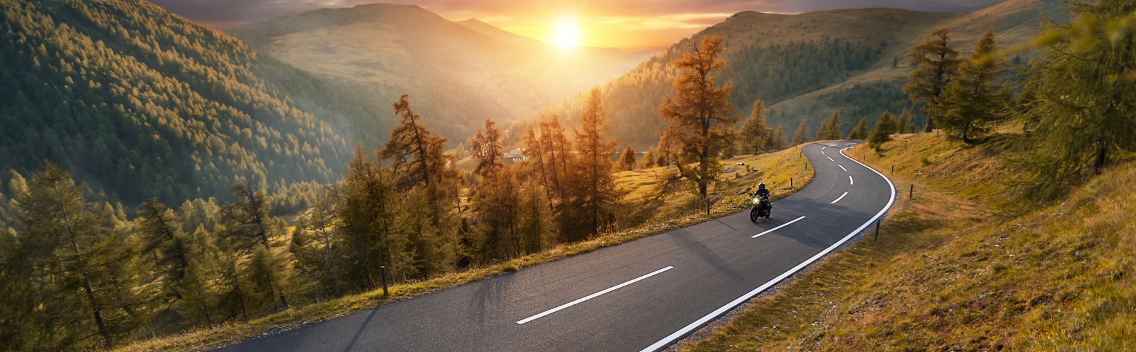 A beautiful valley landscape showing a rising sun and an open road with a single motorcyclist.