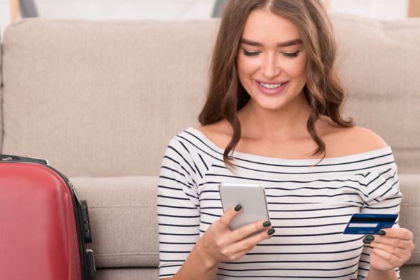 Buying tour online. Girl preparing for vacation, using phone and credit card at home