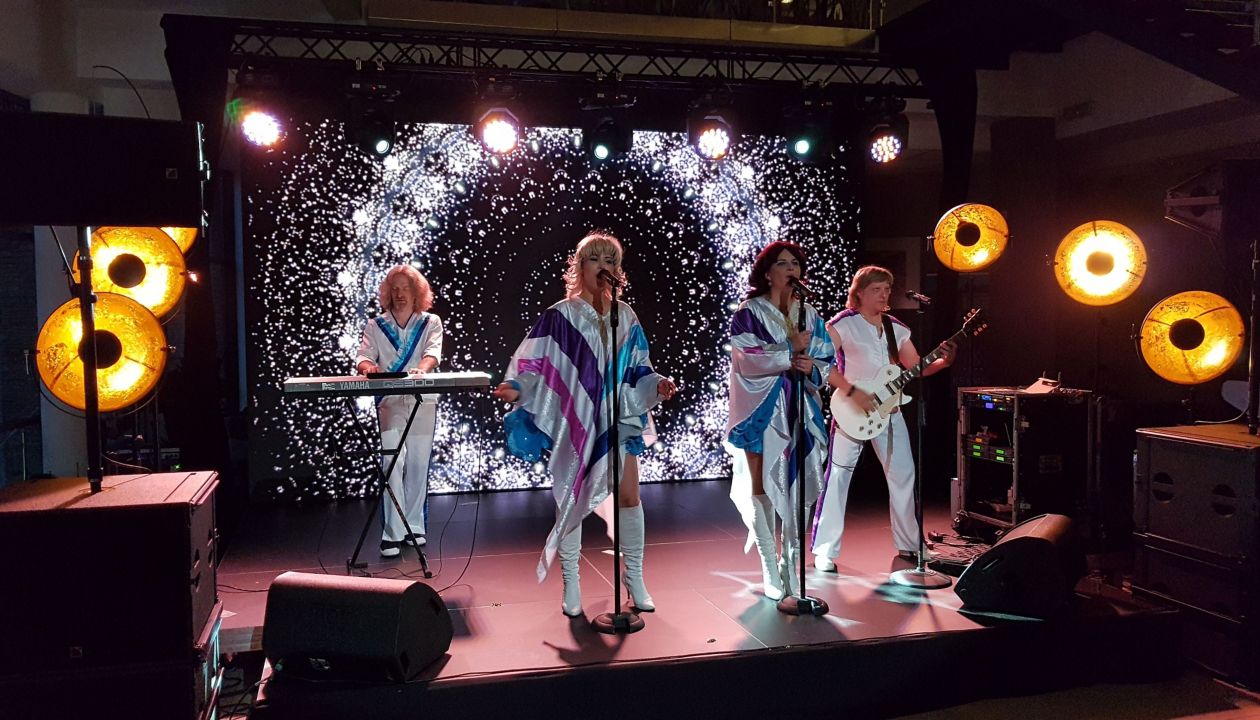 Trzech mężczyzn śpiewających z entuzjazmem karaoke z piosenkami ABBA podczas rejsu Stena Line, w radosnej atmosferze imprezy na pokładzie.