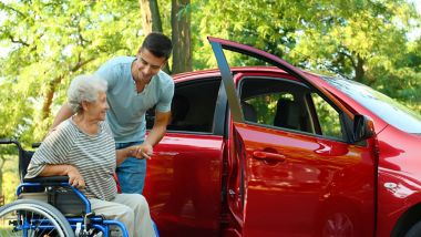 Ung mann som hjelper bevegelseshemmet eldre kvinne i rullestol med å komme inn i en bil