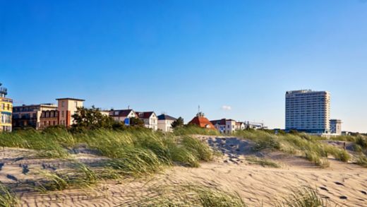  En strand med sand, gräs och träd nära Rostock i Tyskland med byggnader, hus och ett högt hotell.