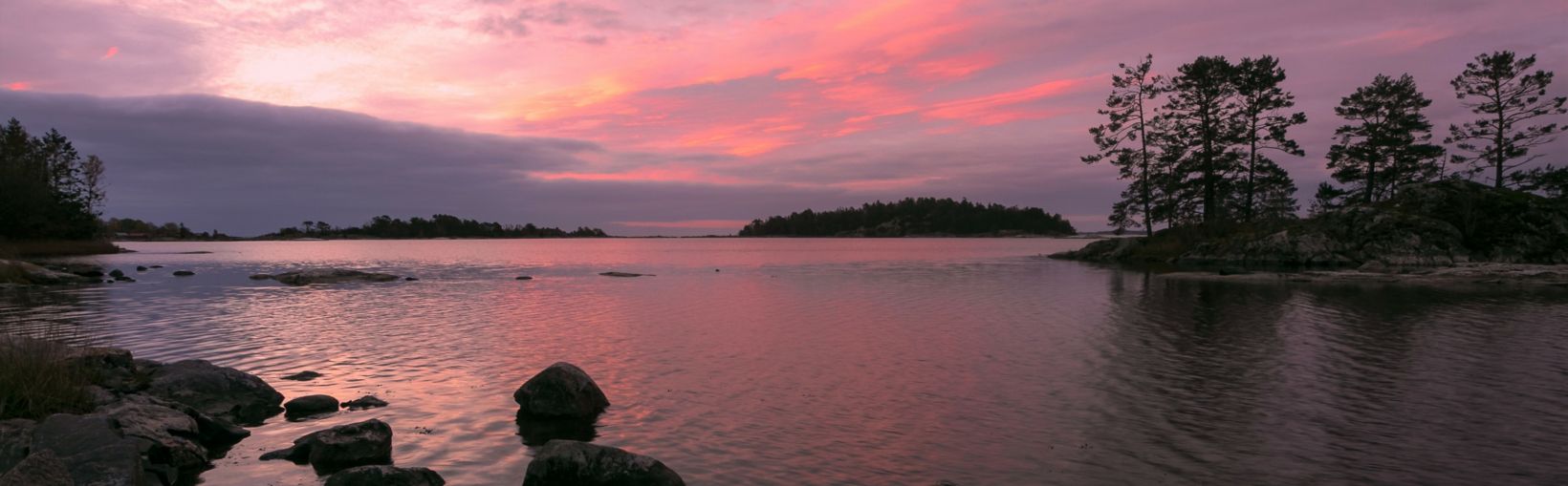 Udsigt over den stenede kyst af en sø i Västerviks skærgård med øer dækket af træer under en lyserød solnedgang
