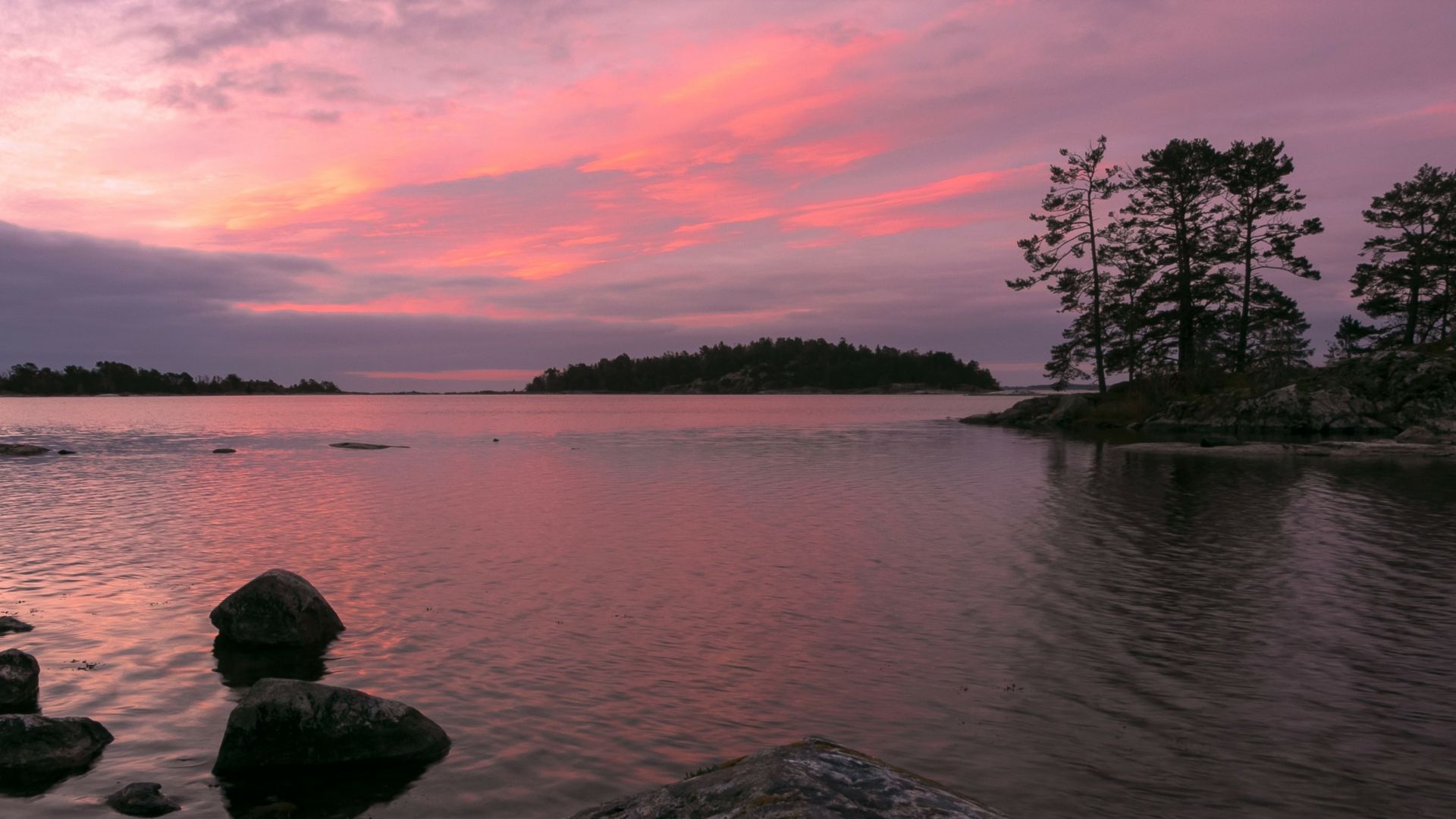 View of the rocky shore of a lake in the Vastervik archipelago with tree covered islands under a pink sunset