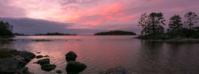 View of the rocky shore of a lake in the Vastervik archipelago with tree covered islands under a pink sunset