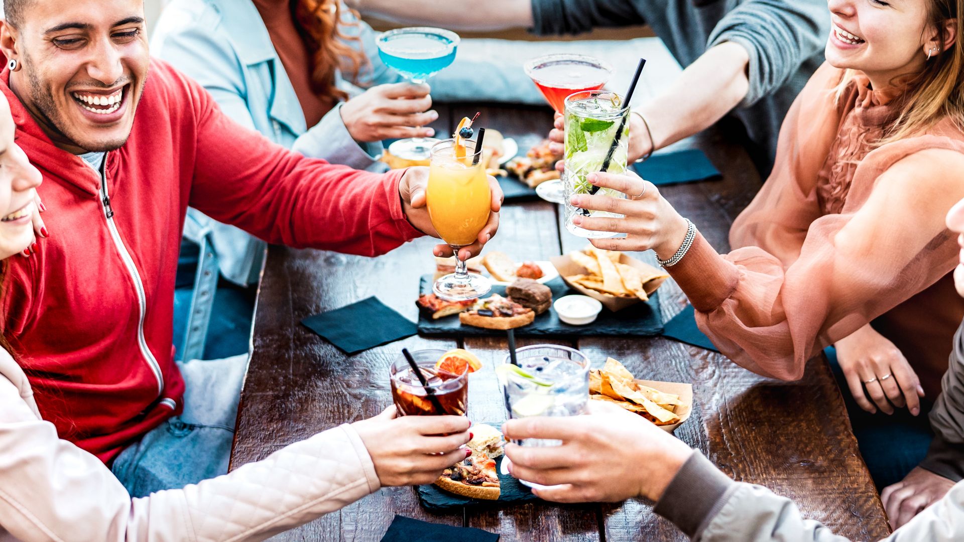 Young people holding multicolored drinks - Trendy fashion friends having fun together toasting cocktails at happy hour - Social gathering life style concept on vivid filter with focus on mid glasses