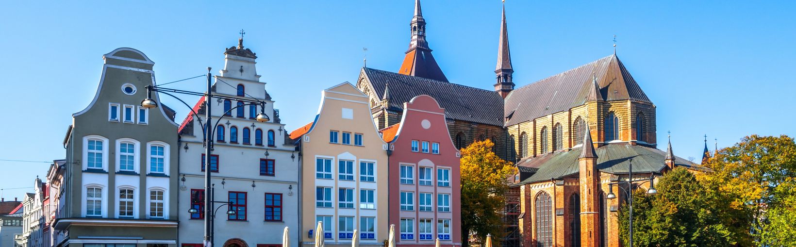 Vue d'un grand nombre de personnes mangeant et buvant à l'extérieur des bâtiments traditionnels multicolores sur le côté du Neuer Markt à Rostock par une journée ensoleillée. L'église St Mary se dresse sur le côté, avec ses multiples flèches et ses grands
