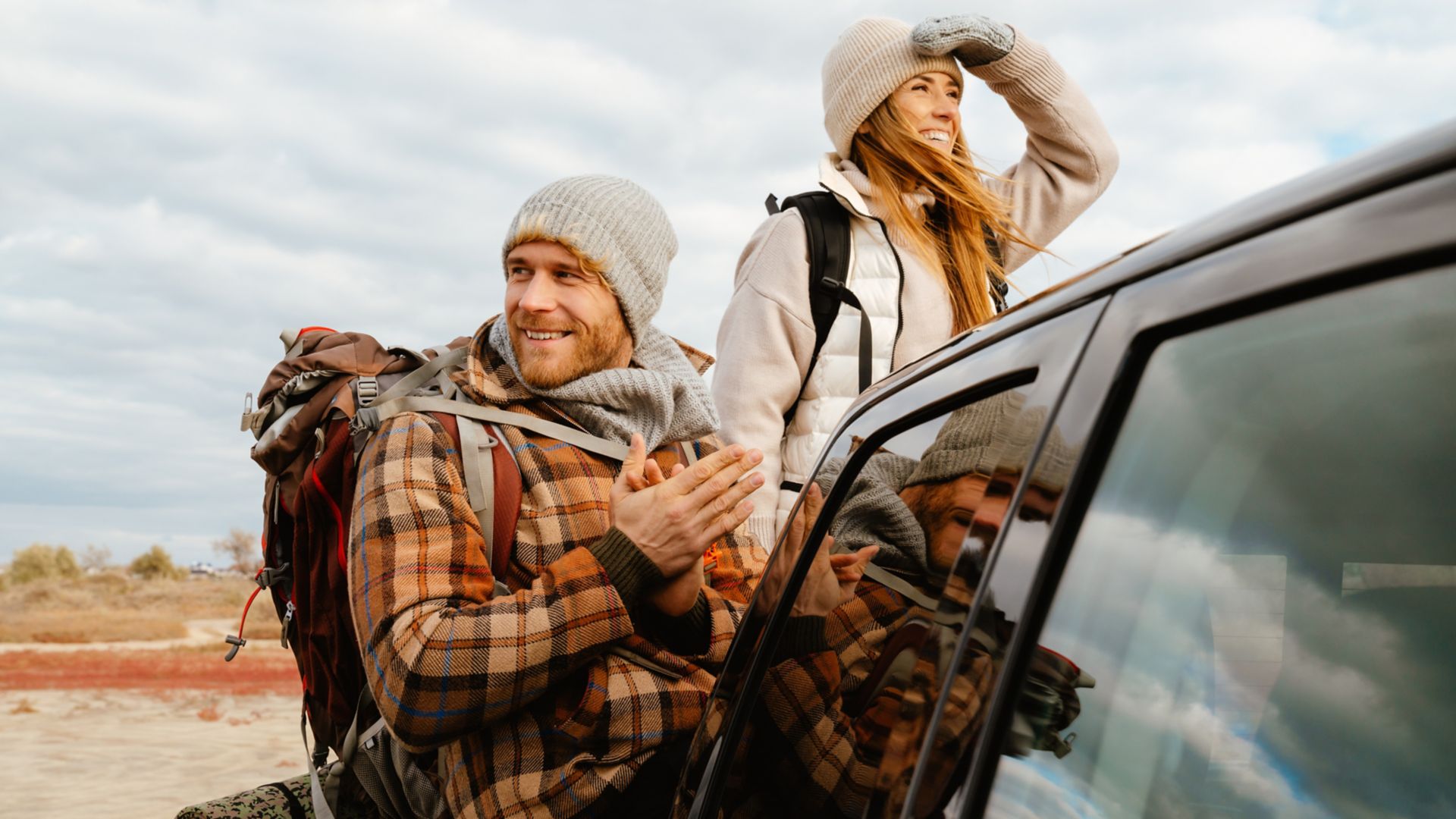 A happy couple in warm clothes and backpacks stand behind their car on an autumn day.