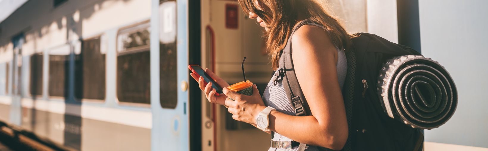 Spoorwegthema. Mooie jonge vrouw met een rugzak gebruikt haar telefoon terwijl ze op het perron bij de trein staat. Goedkope reis.