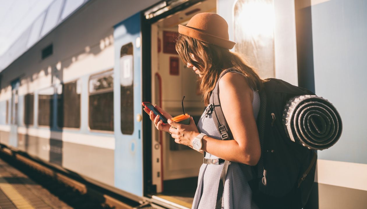 Railroad theme. Beautiful young woman with a backpack uses the phone while standing near the railroad train on the platform. Cheap travel.