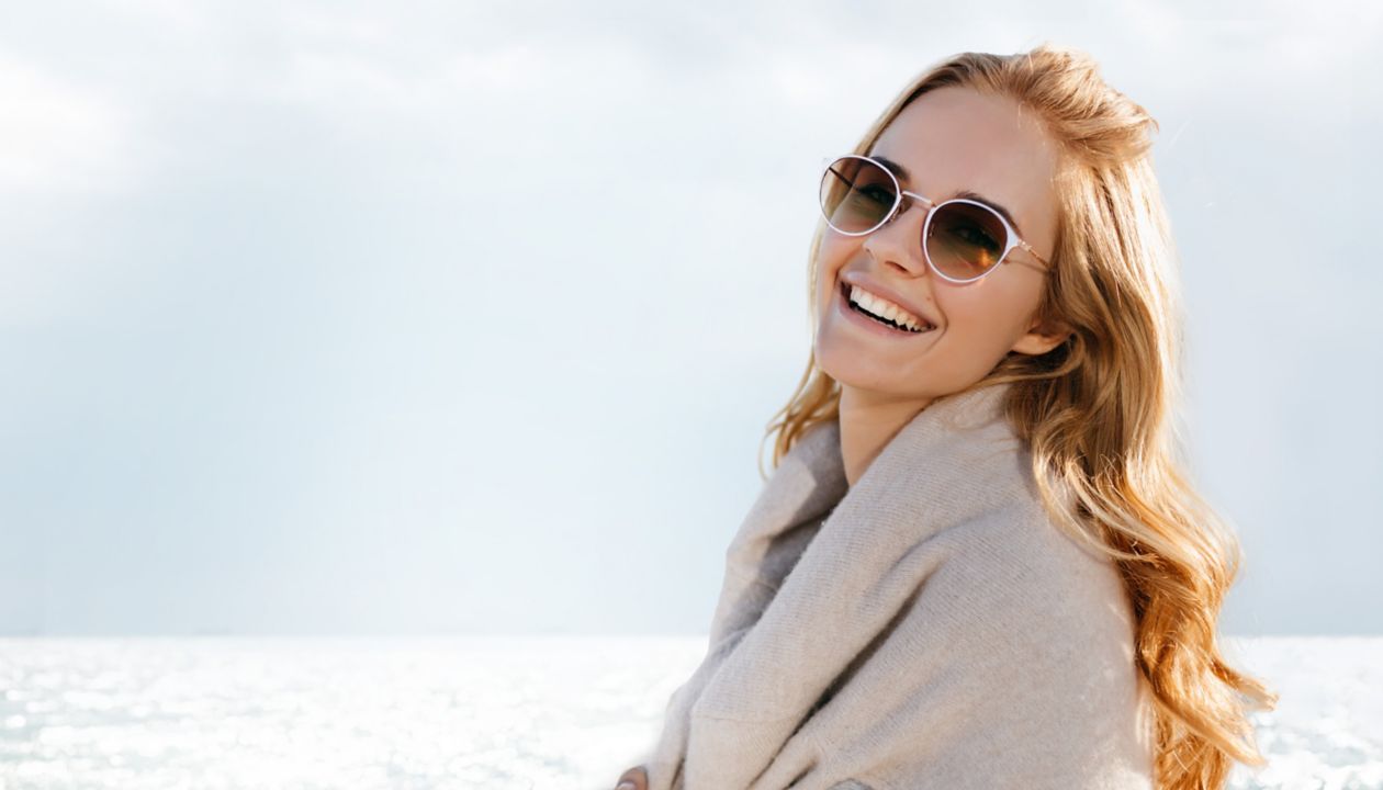 Ruskeita aurinkolaseja käyttävä nainen nauraa ja katsoo kameraan meri taustallaan