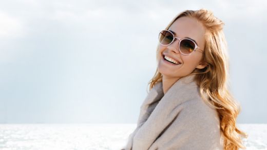 Kvinna i bruna solglasögon skrattar och tittar mot kameran med havet som bakgrund