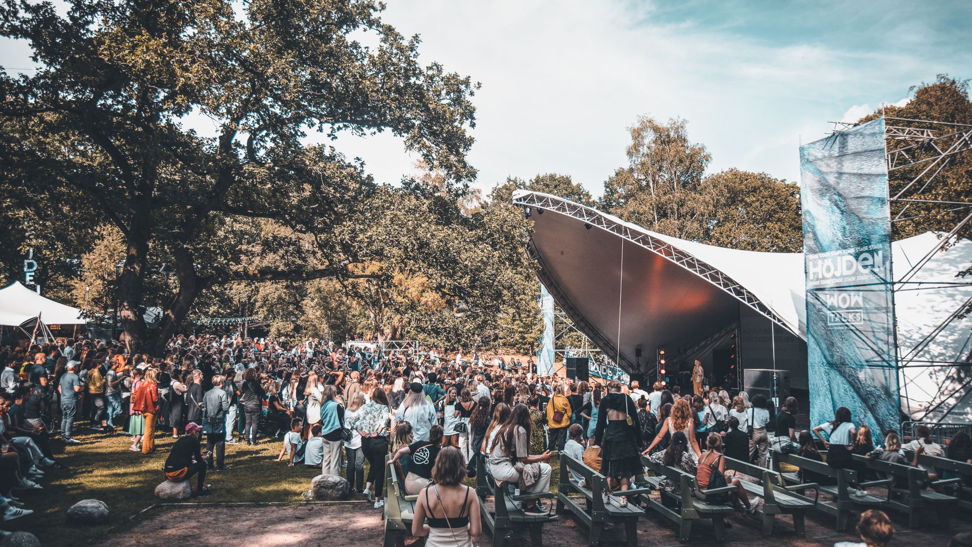 En skare af mennesker, de fleste stående, men nogle siddende, ser en optræden af en person på en scene under et stort lærredstag på Way Out West-festivalen i Göteborg en sommerdag.