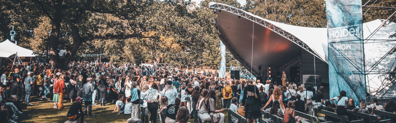 En skare af mennesker, de fleste stående, men nogle siddende, ser en optræden af en person på en scene under et stort lærredstag på Way Out West-festivalen i Göteborg en sommerdag.