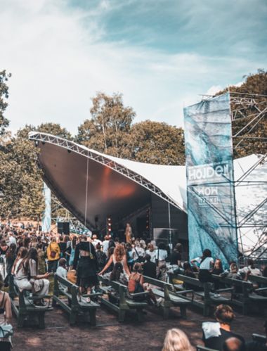 En skare af mennesker, de fleste stående, men nogle siddende, ser en optræden af en person på en scene under et stort lærredstag på Way Out West-festivalen i Göteborg en sommerdag.