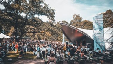 En skare af mennesker, de fleste stående, men nogle siddende, ser en optræden af en person på en scene under et stort lærredstag på Way Out West-festivalen i Göteborg en sommerdag.