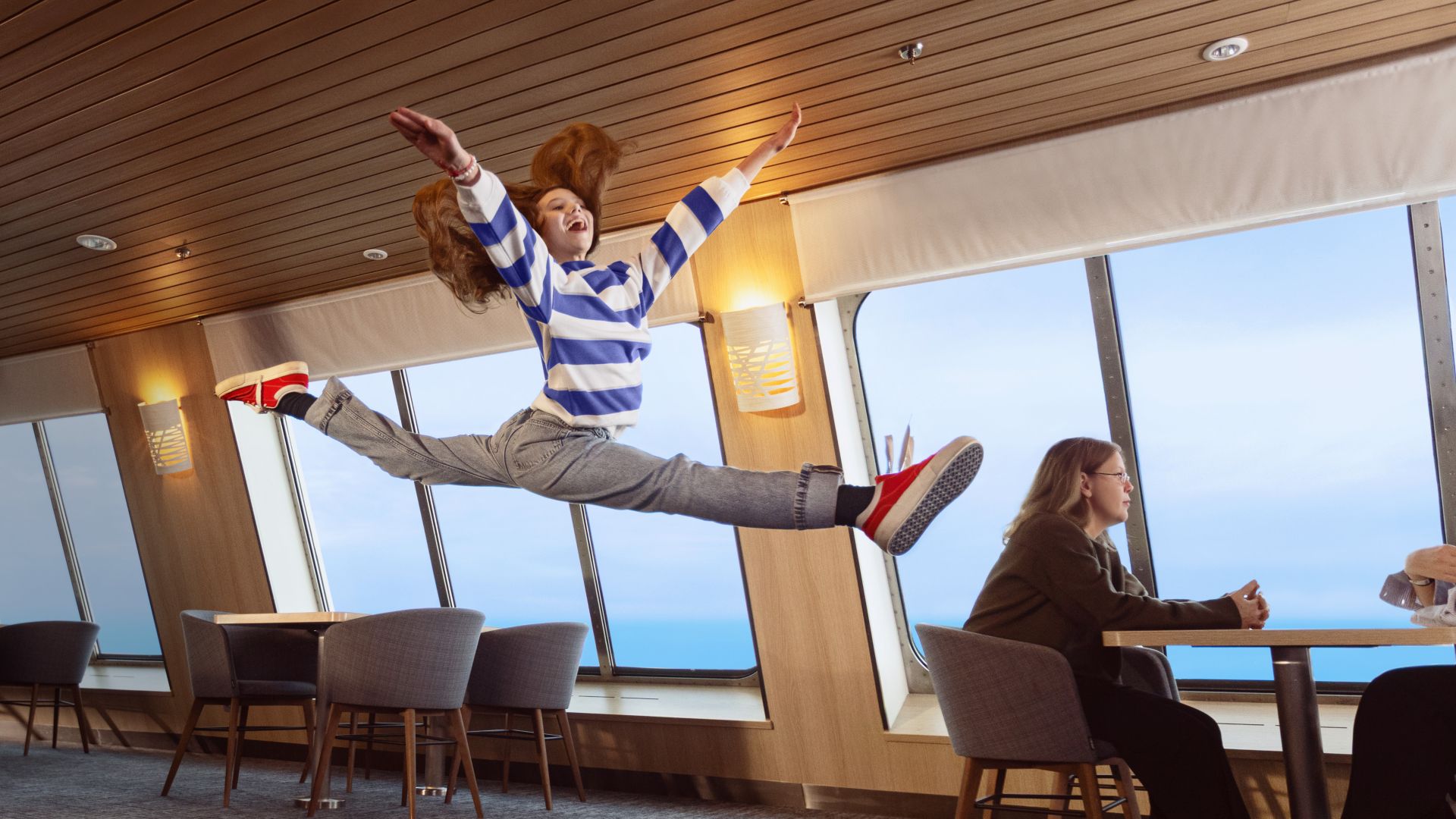A young girl, with long red hair and wearing a stripy jumper, jumps in the air in the Stena Plus lounge.