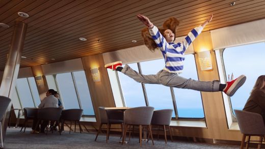 A young girl, with long red hair and wearing a stripy jumper, jumps in the air in the Stena Plus lounge.