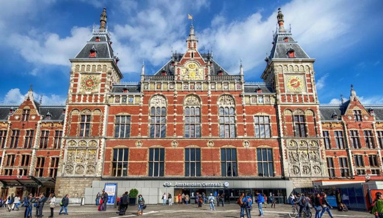Amsterdam Centraal Station with a red-brick facade, ornate towers, clocks, and people walking in front under a blue sky with clouds.