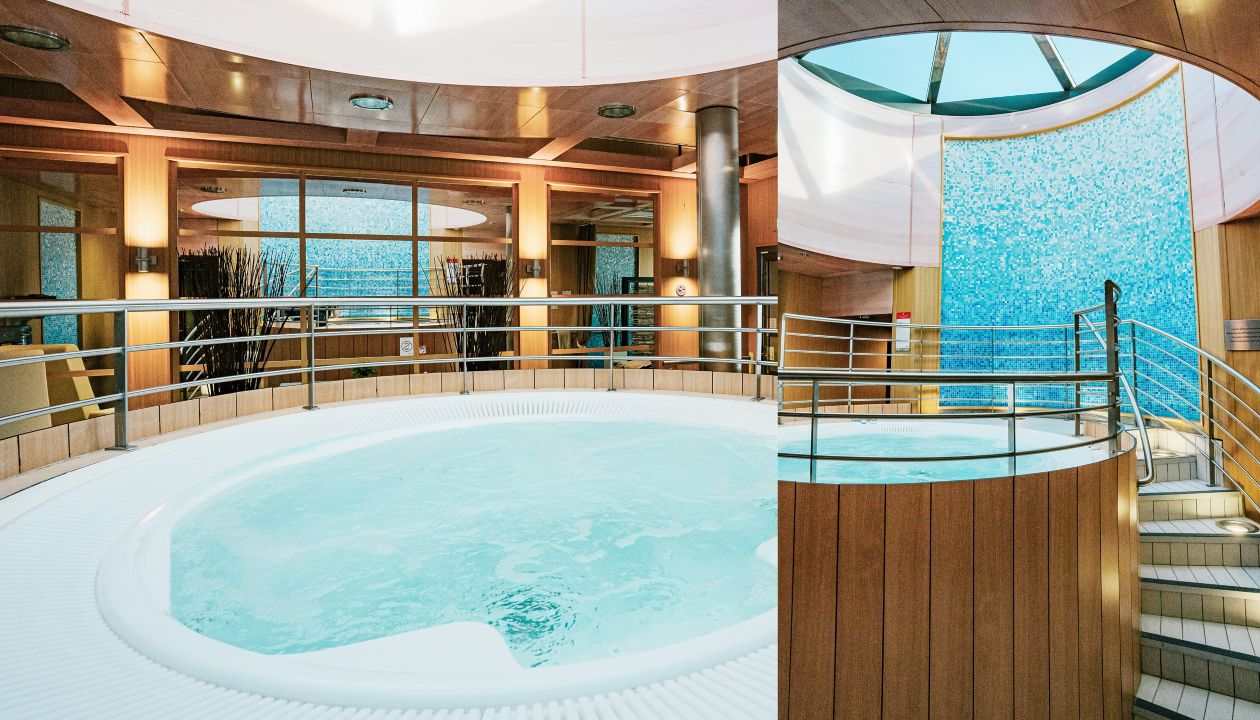 A large circular whirlpool dominates an indoor luxury spa area. Warm wooden paneling and metal railings surround the bubbling pool, creating a modern and inviting atmosphere. Natural light enters through skylights and large windows, highlighting the blue water and textured glass wall. The space appears calm and relaxing, designed for wellness and hydrotherapy experiences.