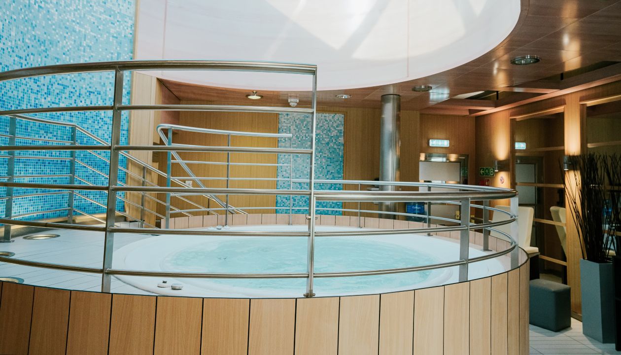 A circular indoor jacuzzi is featured in a contemporary spa setting. The tub is surrounded by wooden paneling and sleek metal railings, giving it a clean, modern look. Blue mosaic tiles and warm ambient lighting enhance the relaxing atmosphere. A large oval skylight above allows natural light to brighten the space, emphasizing the tranquil wellness environment.