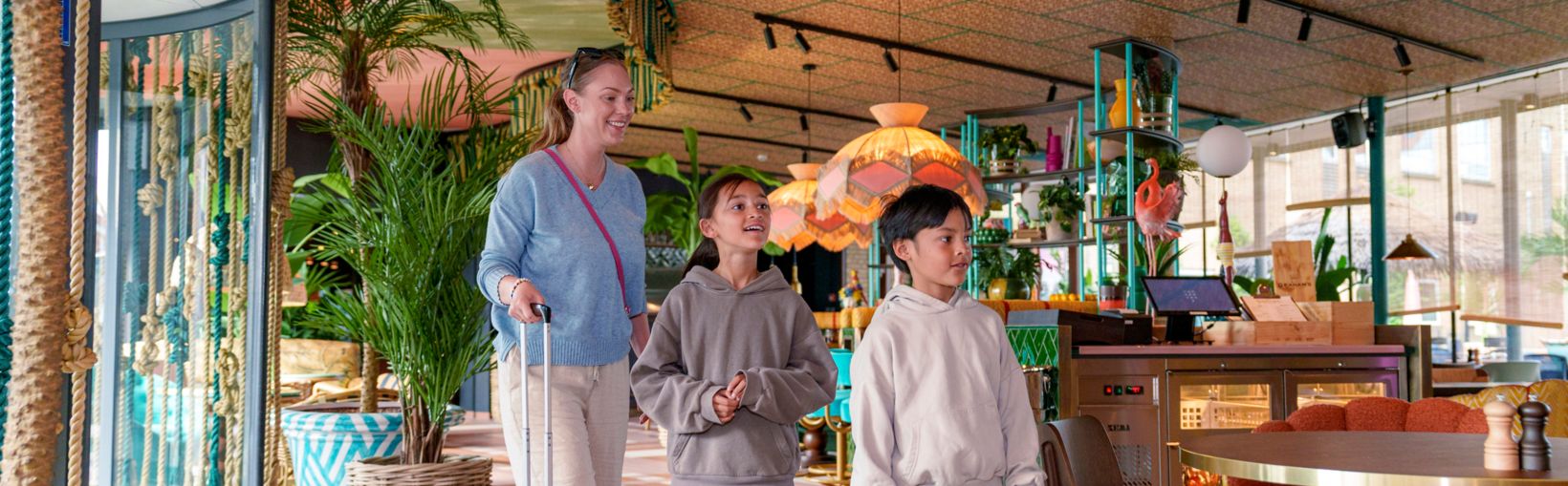 Glimlachende volwassene en twee kinderen komen met bagage aan bij Quality Hotel The Reef in Frederikshavn in een kleurrijke lobby met tropische sfeer.