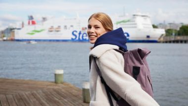 Smiling person wearing a backpack and hoodie standing on a waterfront dock, with a large ferry in the background.