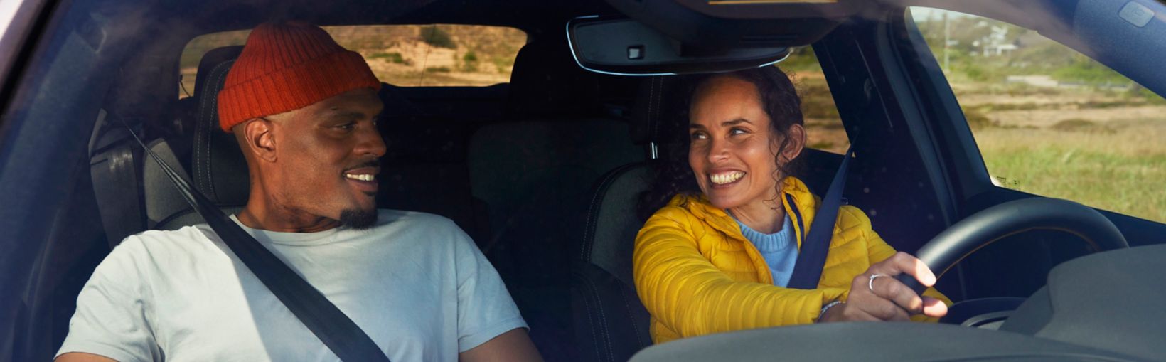 A happy couple are seated in a car, with the female in the driving seat, whilst driving through autumn fields on a bright day.
