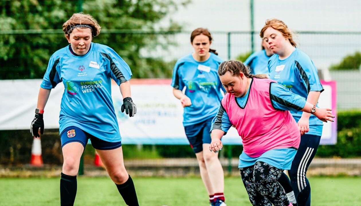 A group of women are actively playing soccer on a green artificial turf field. They are wearing blue sports jerseys, with one player in a pink training bib, and are focused on the soccer ball in play. The background features a fence, trees, and a visible banner. The scene captures movement, teamwork, and a casual, energetic atmosphere. No visible numbers or explicit product branding are present.