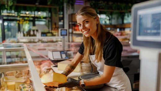 Leende delikatessanställd håller en ostbit bakom en välfylld disk