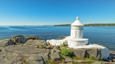 Niewielka biała latarnia morska stoi na surowym, skalistym brzegu nad spokojnym, błękitnym morzem. Ma zwartą, cylindryczną konstrukcję ze stożkowym dachem i znajduje się tuż przy brzegu. Bezchmurne niebo oraz widoczny w oddali ląd po drugiej stronie zatoki tworzą spokojny, nadmorski krajobraz. Pomiędzy skałami rośnie gdzieniegdzie zielona roślinność, która dopełnia naturalny charakter tego miejsca.