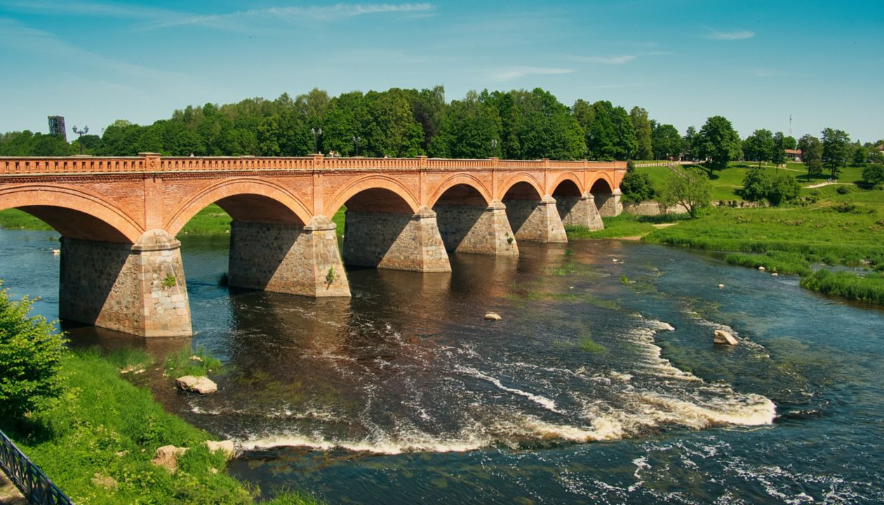 A classic red brick arch bridge spans a gently flowing river, surrounded by lush greenery and trees. The bridge features multiple arches and stone supports, creating a striking architectural focal point. The setting is tranquil, with clear blue skies and vibrant green grass, evoking a peaceful and natural mood. No visible text or numeric values are present in the image.