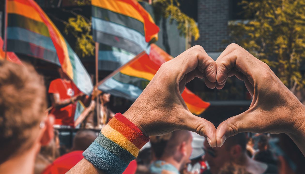 Stützende Hände machen ein Herzzeichen und winken vor einer Regenbogenfahne, die auf einem Wagen einer sommerlichen Gay-Pride-Parade weht