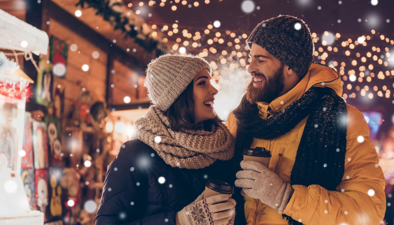 Having fun together at a christmas fairy with snowfall. Young cheerful couple is having a walk with hot drinks, enjoying, dressed warm, looking at each other and laugh, snowflakes all around