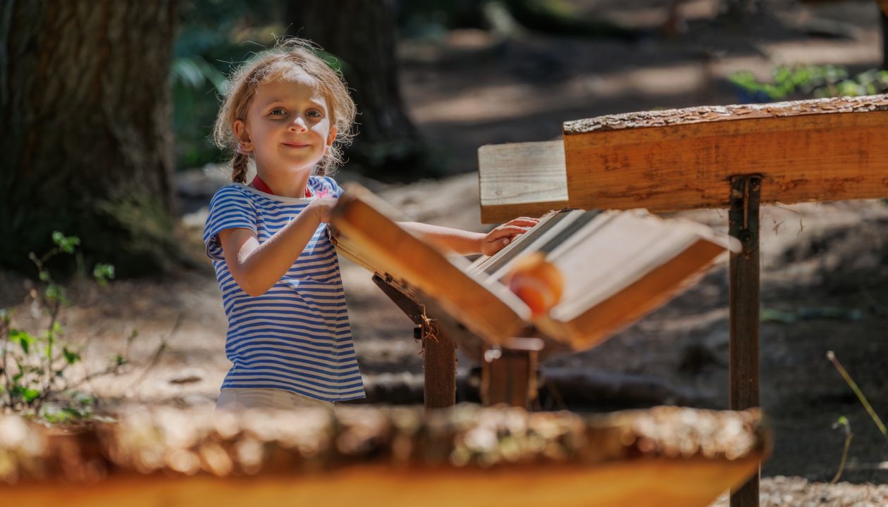 Smiling girl is happily playing with wooden instruments in a forest marble run, fostering creativity and joy while enjoying the beautiful natural environment around her