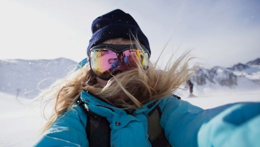 Cool woman makes photo selfie on smartphone on ski slopes in mountains, wears goggles for snow and sun