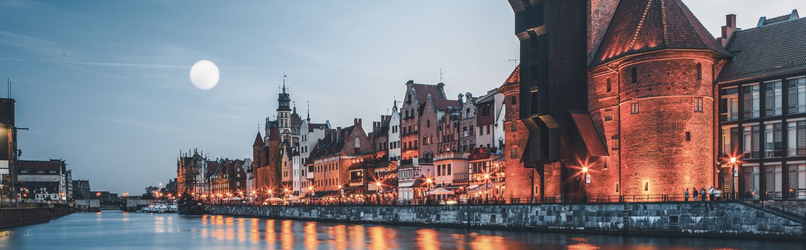 The harbour waterfront of the old town of Gdansk with traditional buildings dimly lit up at dusk.