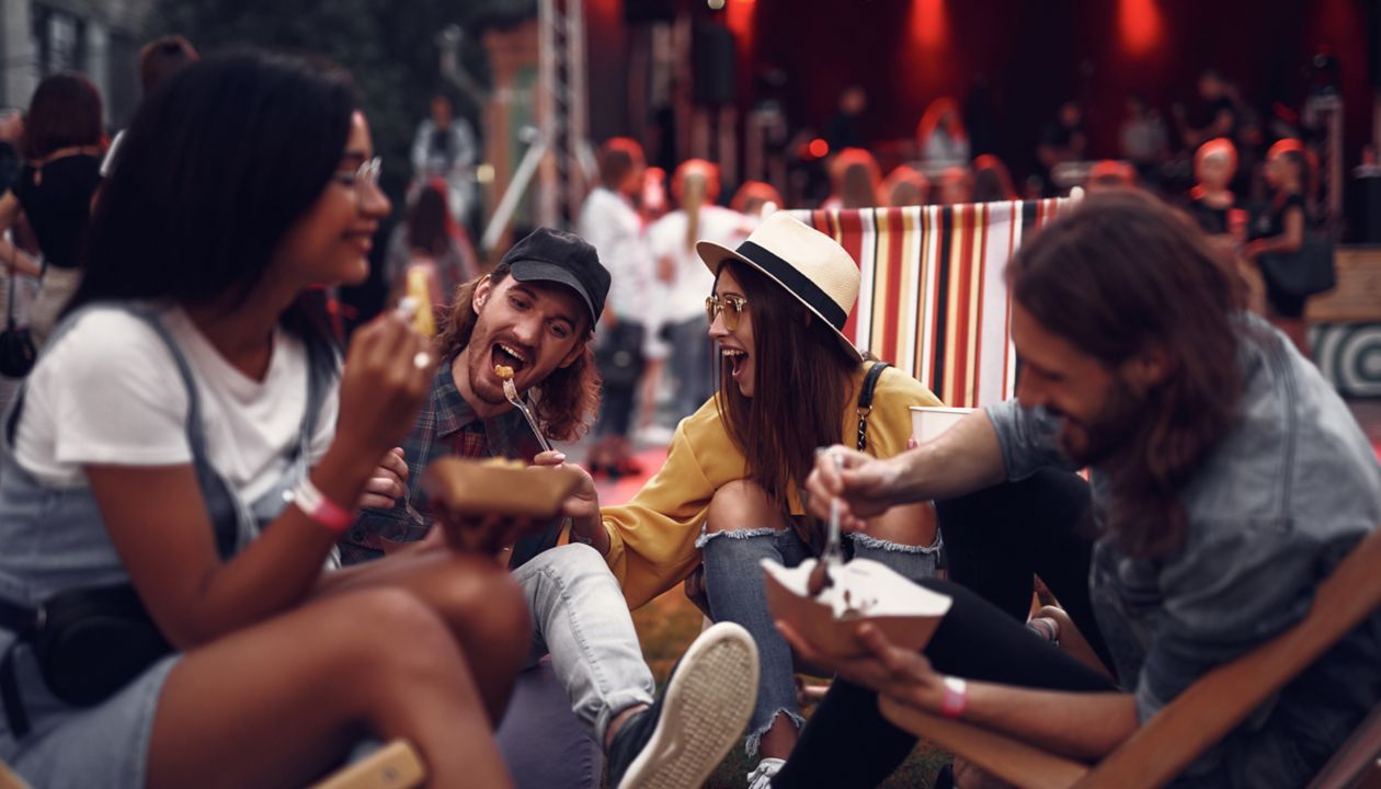 Portrait of cheerful hipster girl in hat feeding boyfriend with fork while sitting on folding chair. Blurred bearded man and girl in glasses eating and smiling