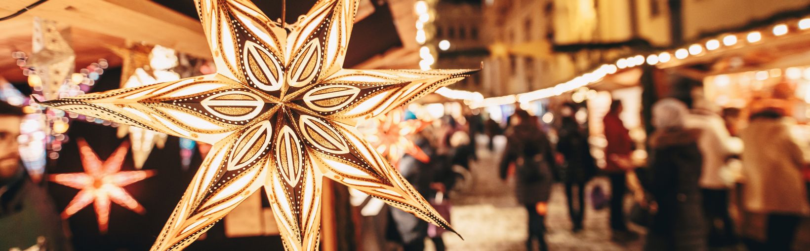 Eine hell erleuchtete Weihnachtsdekoration aus 7 Speichen hängt vor einem Stand auf einem Weihnachtsmarkt.