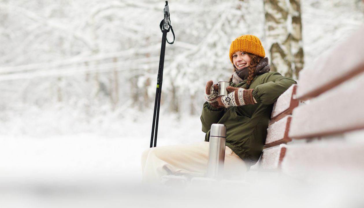 Side view portrait of active young woman smiling at camera while enjoying hot cocoa sitting on bench in beautiful winter forest