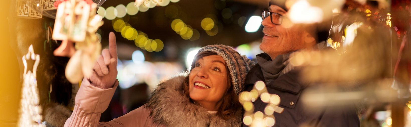 Ett leende par i varma jackor och mössor tittar på juldekorationer vid ett upplyst stånd på en julmarknad.