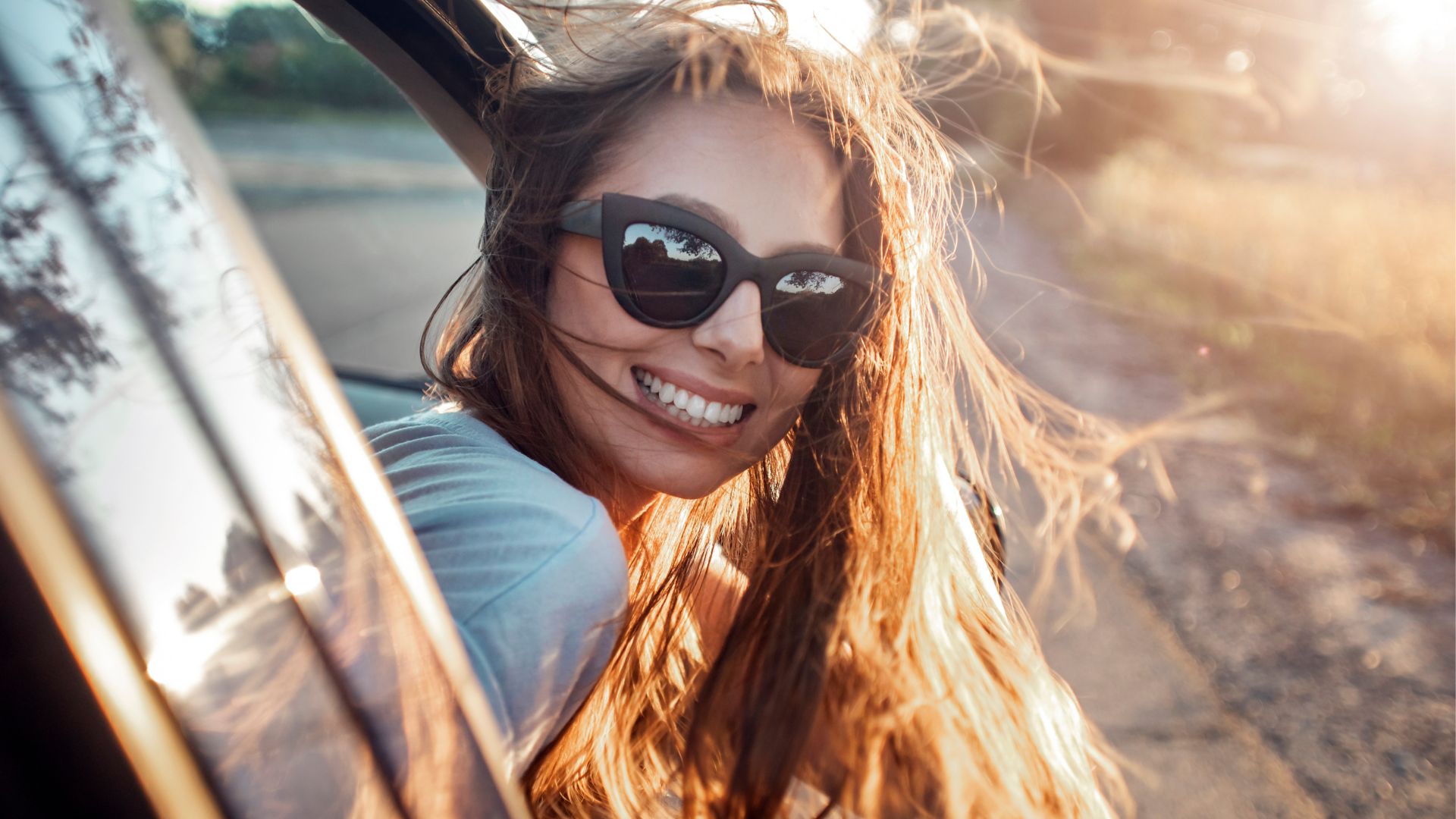 Skaista jauna dāma ar gariem tumšiem matiem un tumšām saulesbrillēm spilgtā saules gaismā izliecas pa novietotas automašīnas logu un skatās uz automašīnas aizmuguri.