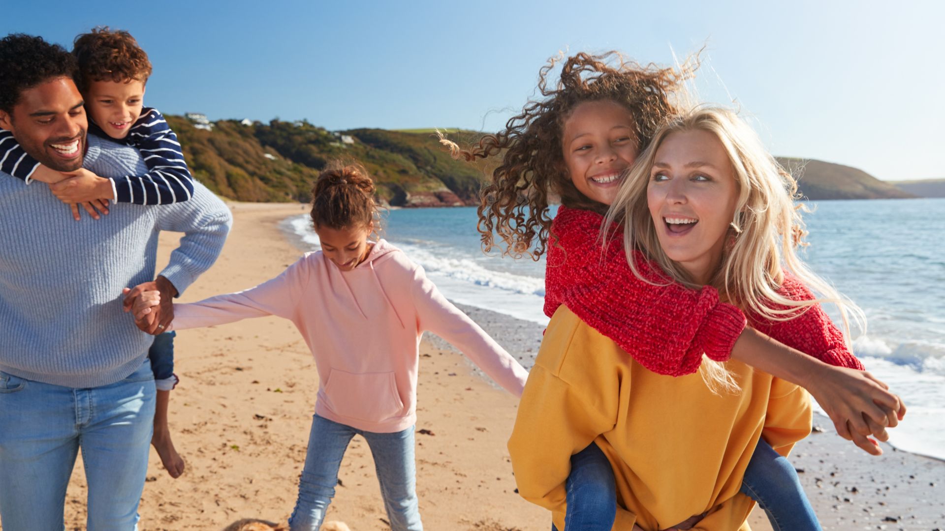 Parents Giving Children Piggybacks As They Walk Along Beach Together