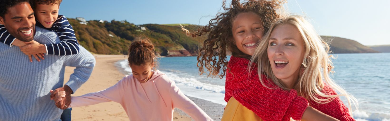 Parents Giving Children Piggybacks As They Walk Along Beach Together