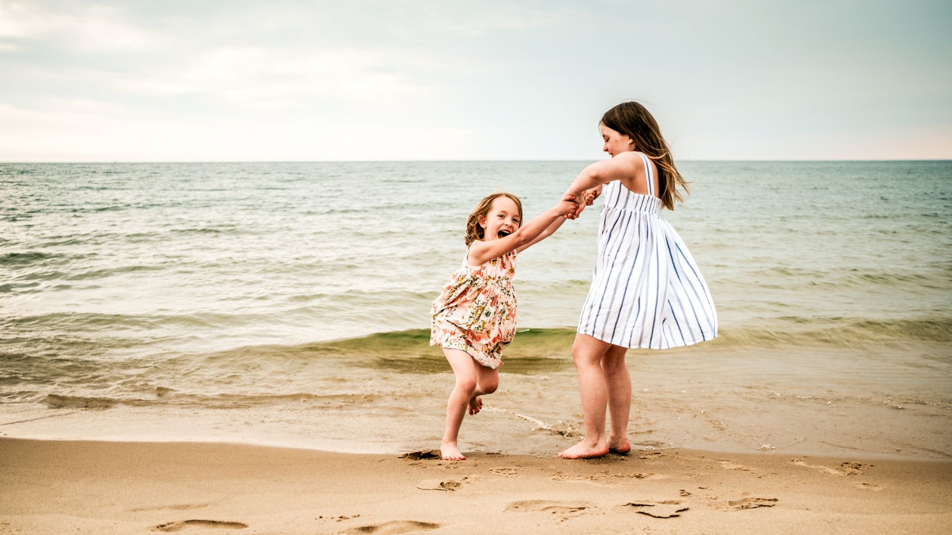 Oudere zus in een wit met blauw gestreepte zomerse jurk duwt haar jongere zus in een bloemmotief zomerse jurk op de schommel op het strand, dicht bij het water, op een bewolkte dag.