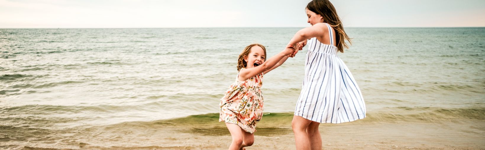 Oudere zus in een wit met blauw gestreepte zomerse jurk duwt haar jongere zus in een bloemmotief zomerse jurk op de schommel op het strand, dicht bij het water, op een bewolkte dag.